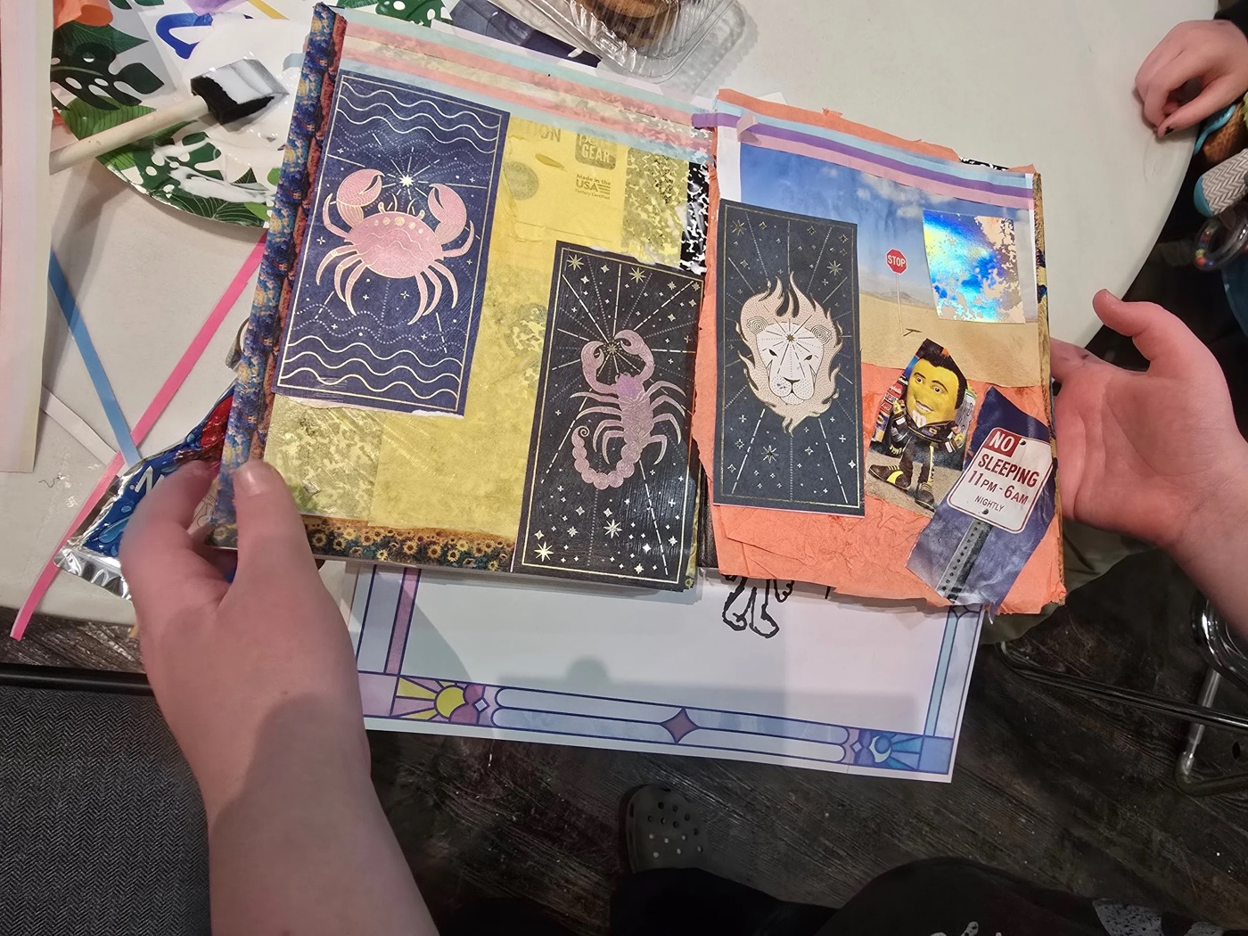 Hands holding a journal spread collaged with zodiac imagery.